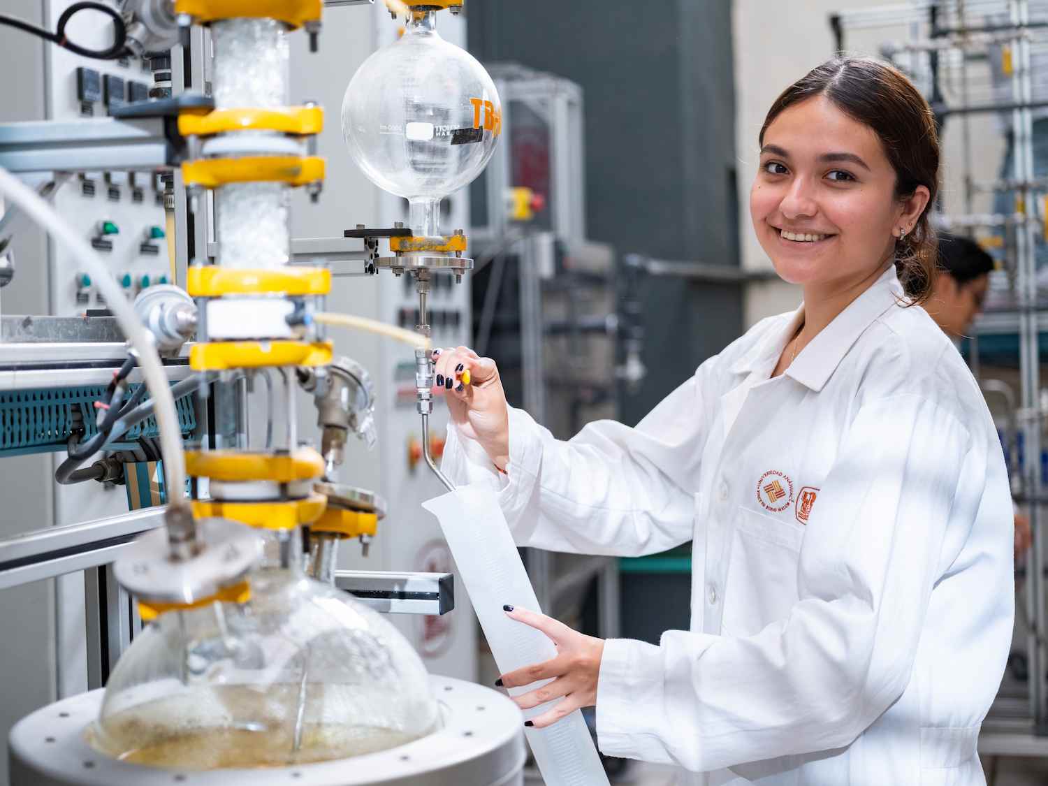 Alumna de ingeniería química sonríe mientras trabaja con equipo de destilación en laboratorio industrial.
